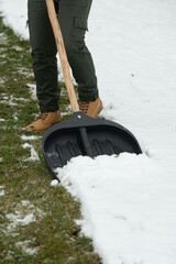 Close-up view of a person wearing work boots and green pants shoveling snow from a lawn. The snow...