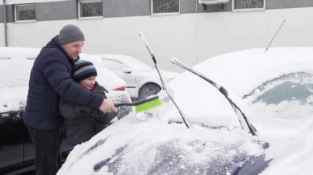 Father and son clean the car from snow with a brush.