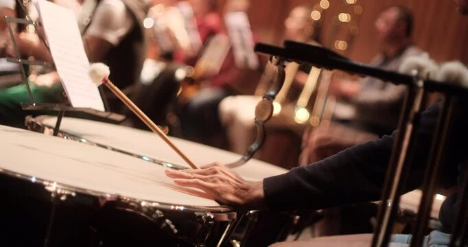 A close-up view of a timpani player with drumsticks poised over the instrument, and sheet music visible on a stand. The blurred background captures other orchestra musicians