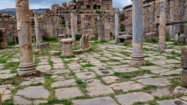 A captivating reveal shot of the Cosinius Market Square and the surviving columns in the ancient Roman settlement of Djemila, Algeria, set against a backdrop of cloudy spring skies