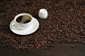 White Coffee Cup with Black Coffee, Creamer, and Scattered Coffee Beans on Wooden Surface