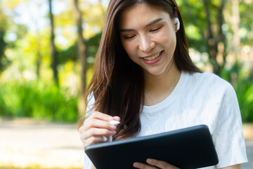 Young Asian female Student in casual clothes sits under the sunlight in a park, using her tablet for research and studying online, e-learning, working on her homework sitting outdoor in a public park.