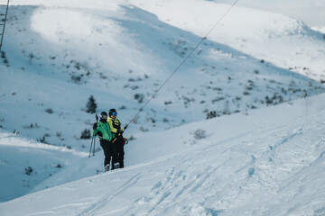 Two skiers ride a rope tow up a snowy mountain slope during a sunny winter day, surrounded by scenic hills. The image captures outdoor adventure and winter sports amidst serene natural beauty.