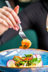 A close-up of a woman's hand holding a fork with a shrimp over a vibrant salad. The dish features greens, radishes, and colorful ingredients, showcasing a fresh and appetizing meal.