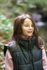 Cute beautiful smiling long-haired girl in glasses and vest stands in the forest. Concept of...