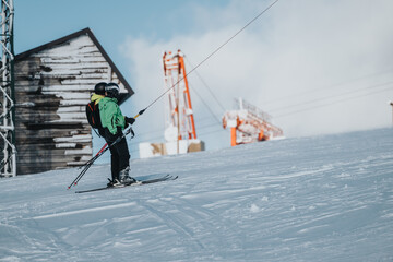 Two skiers ride up a snowy slope on a rope tow lift, wearing warm gear, with a rustic shack and sky...