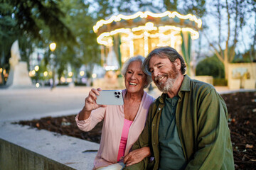 Naklejka premium Happy senior couple taking selfie in amusement park with carousel in background