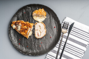Pieces of chicken cutlet Cordon Bleu with bacon and cheese according to a traditional French recipe on a black mica plateon a gray background with napkin and cutlery. Space for text.