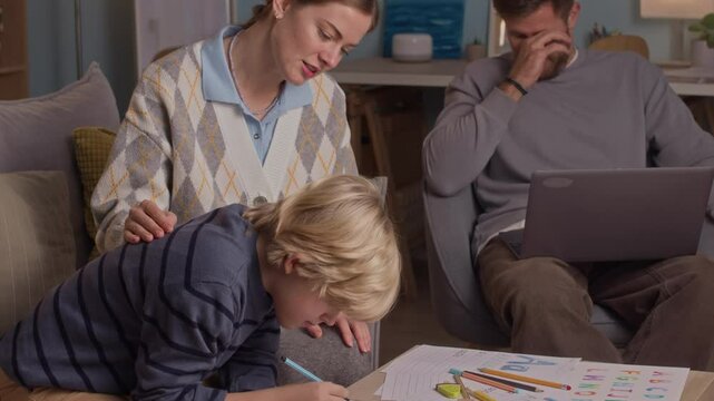 Tilt down shot of young Caucasian mom in smart casualwear helping son with homework while tired dad working on wireless laptop in background