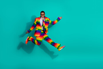 Joyful individual in a rainbow suit jumps energetically on a vibrant turquoise backdrop, celebrating individuality and vibrant energy.