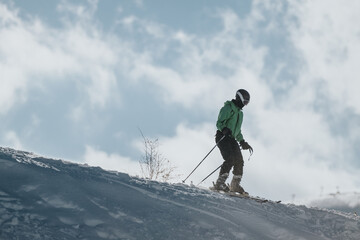 A skier wearing a green jacket and helmet descends a snow-covered slope, enjoying outdoor winter activities under a bright and cloudy sky.