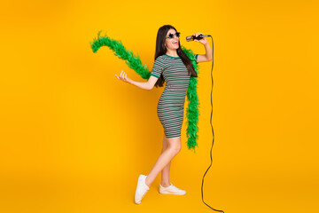 Young woman joyfully singing on microphone with green boa and star sunglasses against a vibrant yellow background