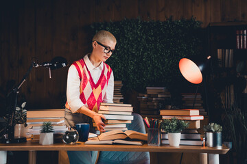 Young woman with short blond hair wearing colorful waistcoat and glasses organizing books at home desk in cozy evening light