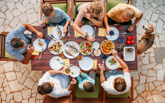 Top view of cheerful family gathered around outdoor dining table enjoying Mediterranean meal with fresh fish, vegetables, and salads, vibrant dishes on rustic terrace, summer vibe, togetherness.