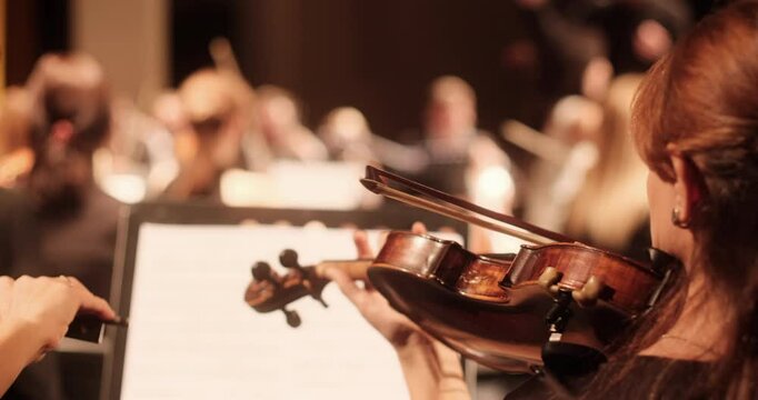 A close-up view of a violin player during a rehearsal of an academic classical orchestra in a softly lit concert hall. The conductor and other musicians are visible in the background