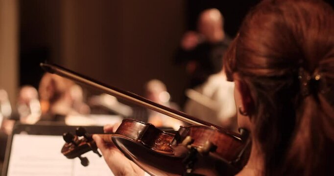 A close-up view of a violin player during a rehearsal of an academic classical orchestra in a softly lit concert hall. The conductor and other musicians are visible in the background