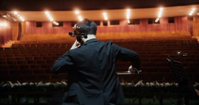 A cellist, captured from behind, rehearses on an empty stage in a grand concert hall. The rows of vacant seats seem to amplify the quiet tension and the anticipation of the upcoming performance