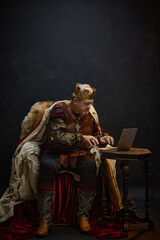 Obraz premium Elderly man as medieval king in crown, sitting on throne and working on laptop against black studio background. Social media marketing, online job. Concept of history, royalty, power, fantasy