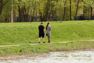 A young guy and his girlfriend are walking in the park along the lake shore. A walk of lovers in the fresh air on a day off. Family leisure in nature