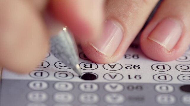 A person is writing on a sheet of paper with a pen. The sheet of paper has a grid of numbers and letters