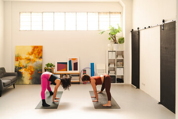 Two women on yoga mats in a bright home gym setting, bending forward while preparing for a deep forward bend pose, surrounded by modern furniture and indoor plants.