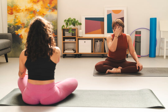 Two women sitting cross-legged on yoga mats, engaging in alternate nostril breathing exercises in a bright home gym with modern decor, indoor plants, and a peaceful ambiance.