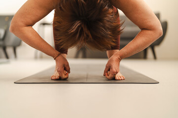 A woman dressed in brown athletic wear holds the plow pose on a black yoga mat in a well-lit contemporary indoor space, with bookshelves and art pieces in the background.
