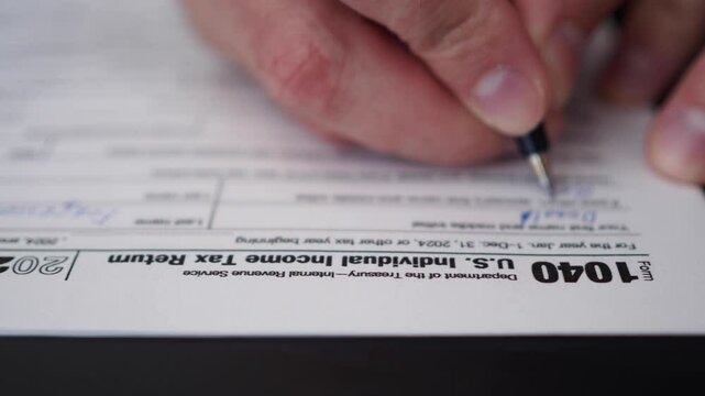 Man Filling In The Tax Form 1040 for 2024 Year. Person Calculates of Planning Budgets Yearly Personal Finance and Fills up US Tax Form 1040 For 2024 Year. Signing 1040 Income Tax Return For 2024 Year.