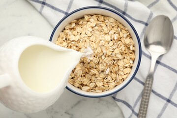 Pouring milk into bowl with oatmeal at white marble table, top view