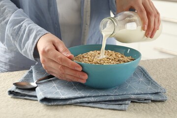 Woman pouring milk into bowl with oatmeal at beige textured table, closeup
