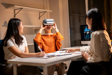 Three women are sitting at a table, one of them wearing a VR headset