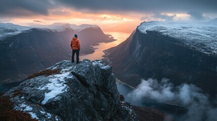 A lone hiker stands on a snowy mountain peak overlooking a valley with a river and dramatic sky at sunset.