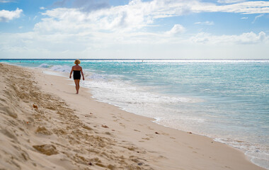 senior woman walks alone along the shoreline of a Caribbean beach with clear sand and deep blue...