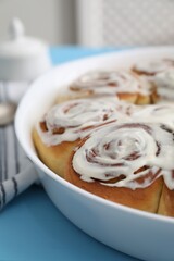 Delicious frosted cinnamon rolls in baking dish on light blue table, closeup