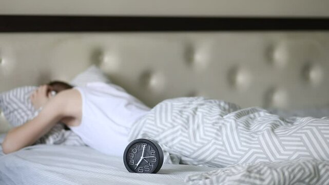 Teenager kid boy lies on bed waking up in morning and gets angry at alarm clock covers his ears with a pillow