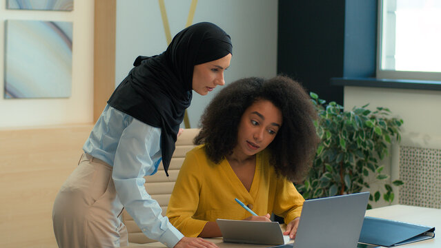 Arabian muslim islamic businesswoman in hijab mentoring new employee African American woman friendly help teach working with laptop in office diverse business women discuss online project write notes