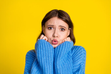 Young woman in blue sweater posing against bright yellow background expressing surprised emotion.