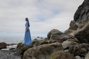 art portrait of woman standing on the rocks near beach and ocean looking over the sea wearing long blue dress