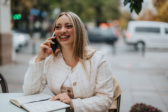 A cheerful woman wearing a white jacket, engaged in a phone call, seated with a notebook in a casual outdoor setting featuring a blurred urban background.