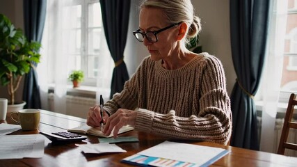 Focused senior woman wearing eyeglasses meticulously reviews financial documents, calculating expenses and managing budget at home, demonstrating responsible financial planning