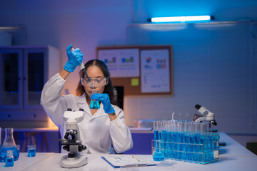 Young scientist wearing lab coat and gloves using micropipette dropping blue liquid into beaker,...
