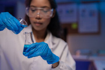 Close-up of scientist wearing lab coat and safety glasses carefully pouring blue chemical liquid...