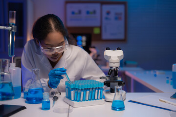 Young scientist wearing a lab coat and gloves, dropping blue liquid into test tubes while...