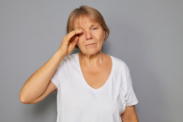 A woman stands against a gray wall, lightly touching her forehead with one hand. She shows a...