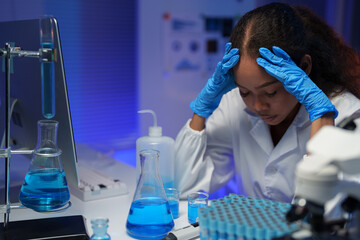 Scientist in a lab coat and gloves sitting at a table in a laboratory, touching her head in stress,...