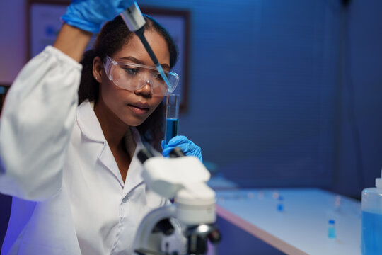 Young female scientist wearing lab coat and protective glasses using pipette to drop blue liquid into test tube during medical research in modern laboratory