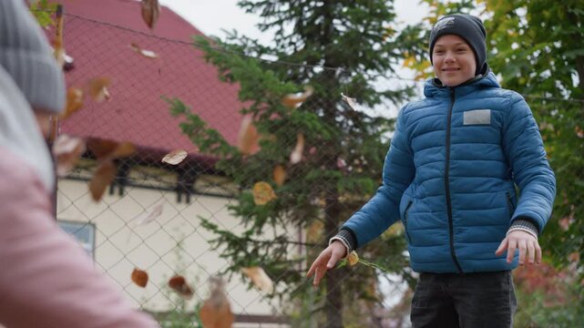 Family fun during autumn as a young boy playfully tosses leaves while his aunt bends down to collect more, both sharing a moment of joy surrounded by colorful foliage and a rustic backdrop