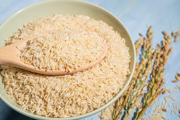 A bowl of brown-rice is placed on a table next to a spoonful and ears of rice.Rice is spread out in the bowl and the spoon is full of raw-rice and sitting on top of it