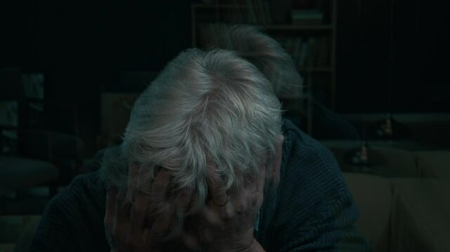 Low key shot of elderly Caucasian man sitting in dark living room covering his face with hands having anxiety attack at home and feeling overwhelmed