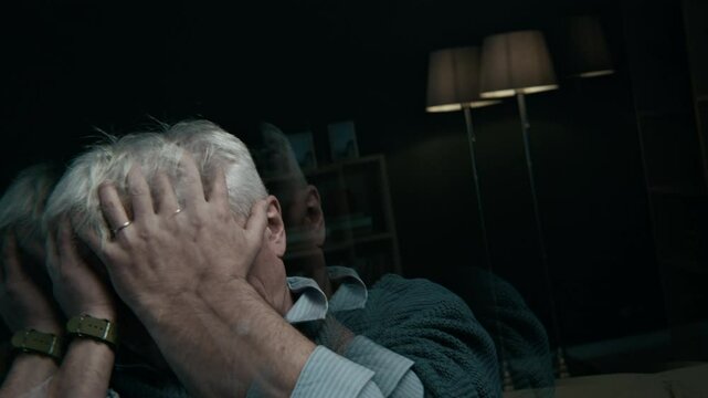 Low key shot of elderly Caucasian man with grey hair rubbing his face feeling anxious and disoriented staying alone at home in dark living room